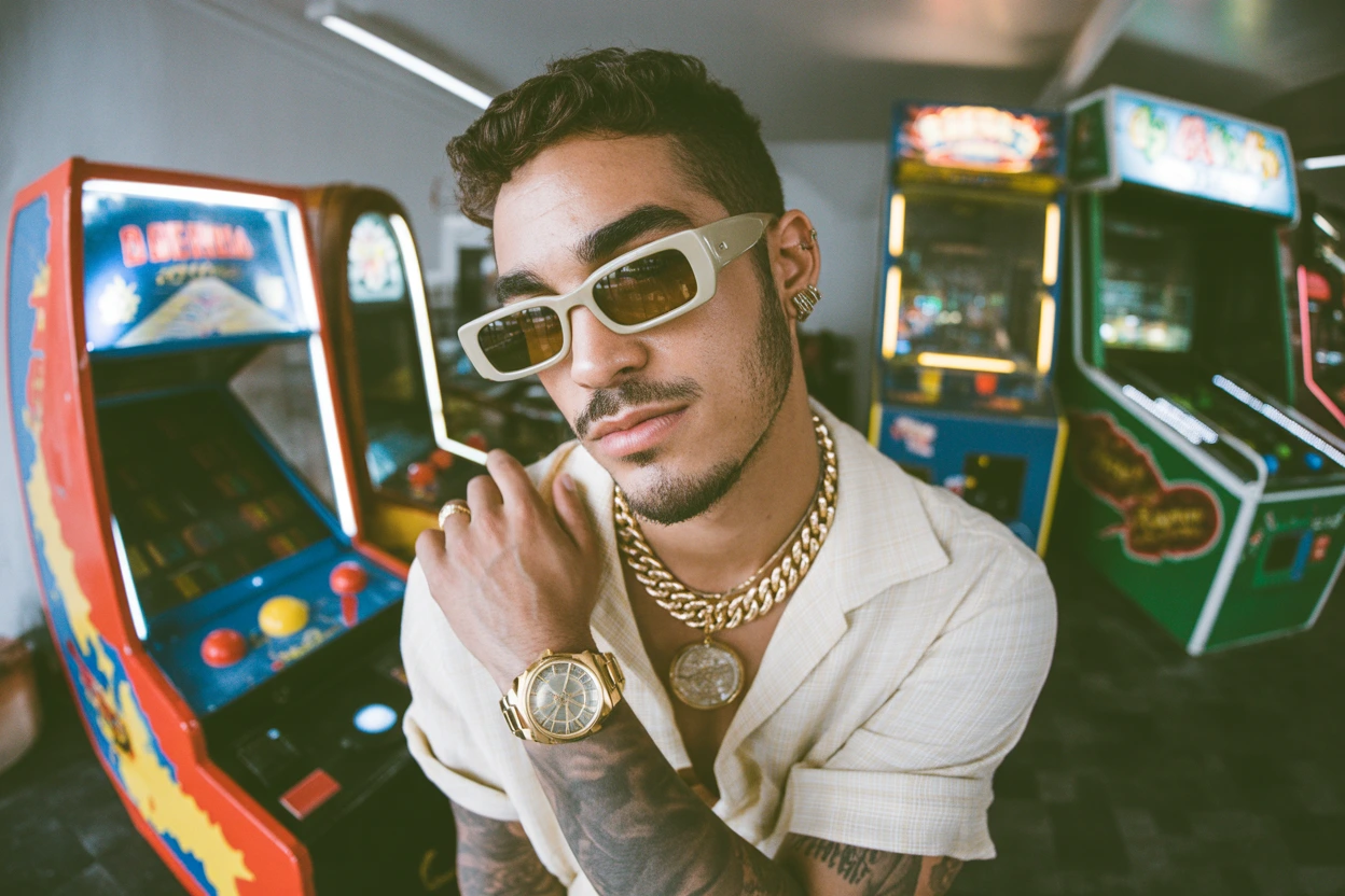 مرد جوانی با استایل رترو، عینک آفتابی، و زنجیر و ساعت طلا که در یک سالن بازی با دستگاههای آرکید قدیمی عکس گرفته است